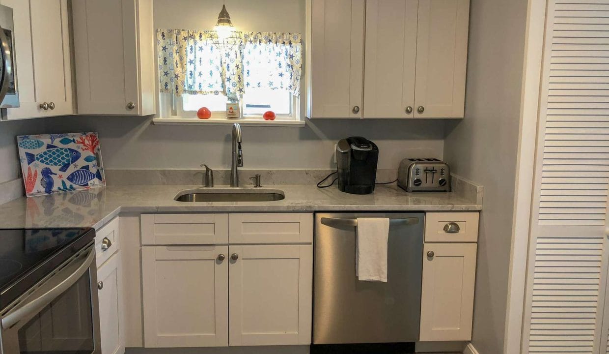 A modern kitchen with white cabinets, stainless steel appliances, a Keurig on the counter, and a towel hanging from the dishwasher. A small window above the sink has floral-patterned curtains.