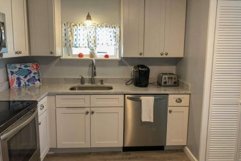 A modern kitchen with white cabinets, stainless steel appliances, a Keurig on the counter, and a towel hanging from the dishwasher. A small window above the sink has floral-patterned curtains.