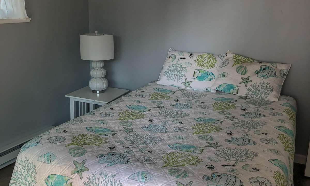 A neatly made bed with sea-themed bedding, featuring fish and coral designs, in a gray-walled room. A white nightstand with a lamp is beside the bed.