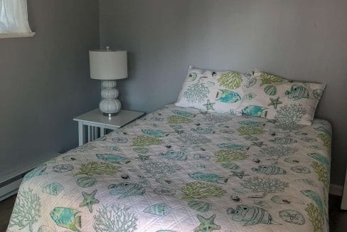 A neatly made bed with sea-themed bedding, featuring fish and coral designs, in a gray-walled room. A white nightstand with a lamp is beside the bed.