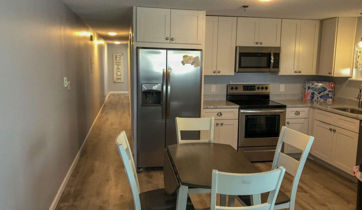 Modern kitchen with stainless steel appliances, light gray cabinets, and a wooden dining table with four chairs. A long hallway leads to other rooms. Walls are painted light gray.