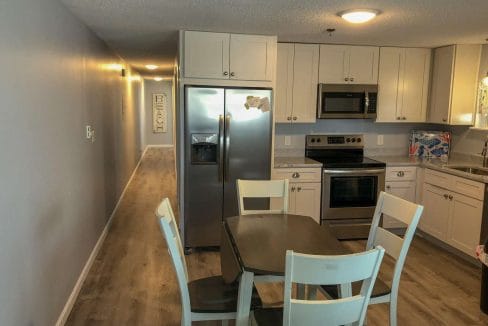 Modern kitchen with stainless steel appliances, light gray cabinets, and a wooden dining table with four chairs. A long hallway leads to other rooms. Walls are painted light gray.