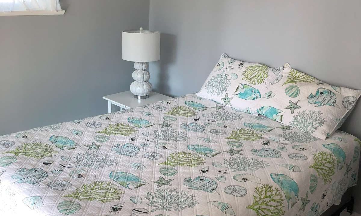 Bedroom with a quilted bedspread featuring sea life patterns, a white side table, matching lamp, and a small window with sheer curtains on a gray-painted wall.