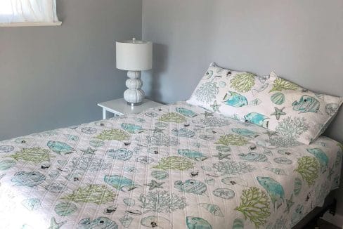 Bedroom with a quilted bedspread featuring sea life patterns, a white side table, matching lamp, and a small window with sheer curtains on a gray-painted wall.