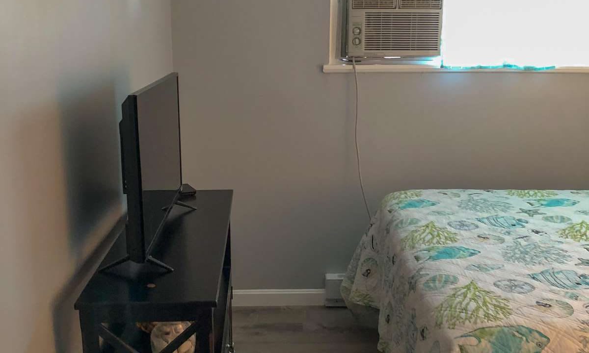 A small bedroom with a bed covered in a patterned quilt. A TV sits on a stand next to the bed. An air conditioning unit is installed in the wall above the bed.