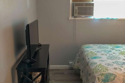 A small bedroom with a bed covered in a patterned quilt. A TV sits on a stand next to the bed. An air conditioning unit is installed in the wall above the bed.