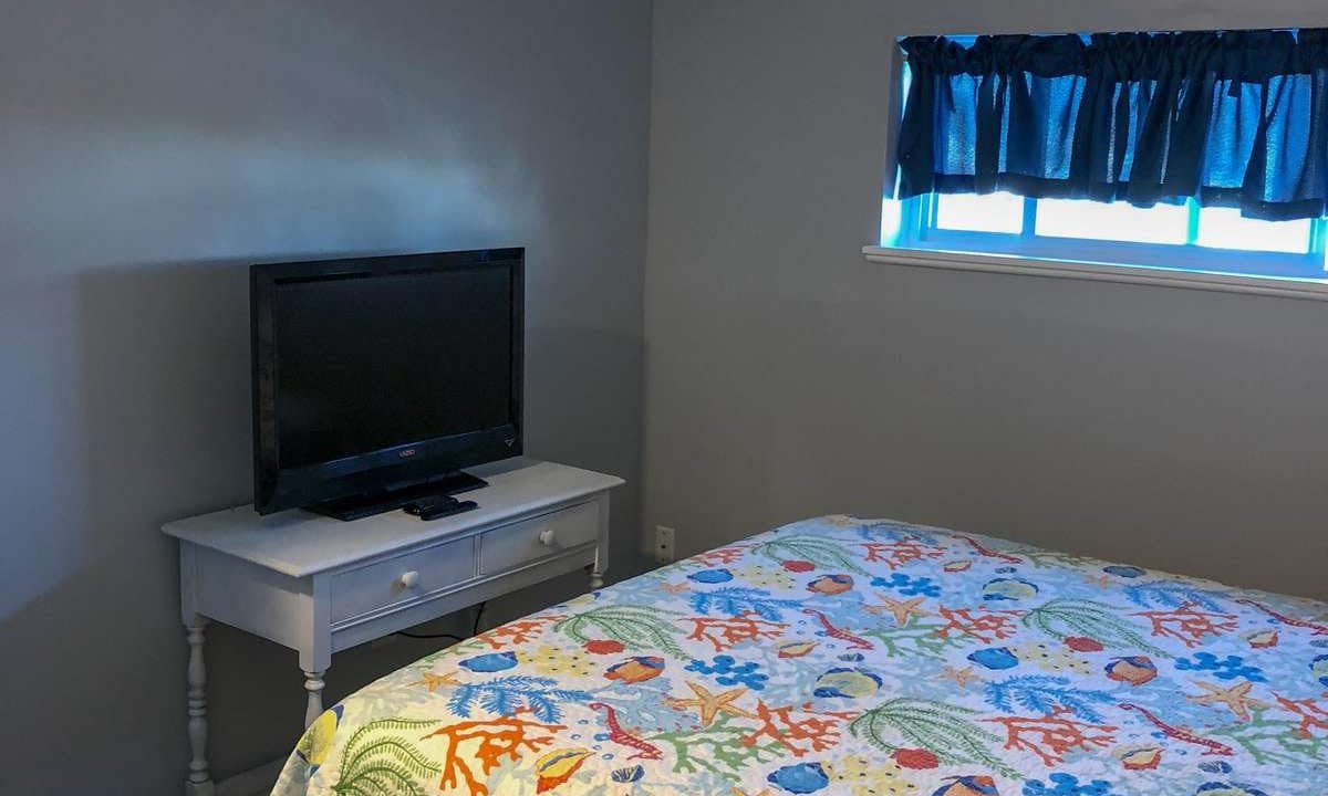 Bedroom with a colorful quilt on the bed, a TV on a small white table, and a window with a dark curtain.