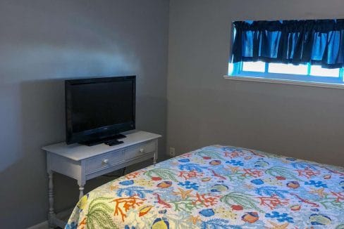 Bedroom with a colorful quilt on the bed, a TV on a small white table, and a window with a dark curtain.