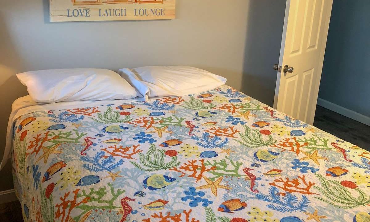 A bed with a colorful ocean-themed quilt, two pillows, and a beach-themed wall art above the headboard. An open door is visible to the right.