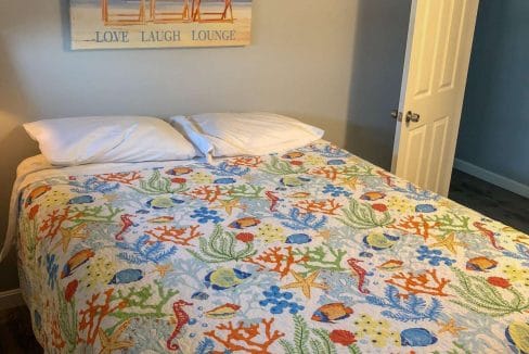 A bed with a colorful ocean-themed quilt, two pillows, and a beach-themed wall art above the headboard. An open door is visible to the right.