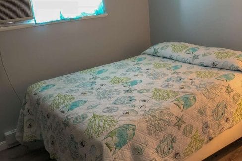 A bed with a marine-themed quilt is in a room with gray walls, a wood floor, and a window with an air conditioner.