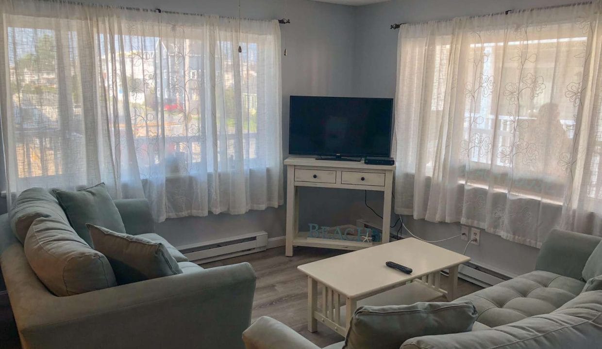 Living room with light green sofas, a white coffee table, a TV on a stand, large windows with sheer curtains, and a ceiling fan. Wood flooring and light gray walls.