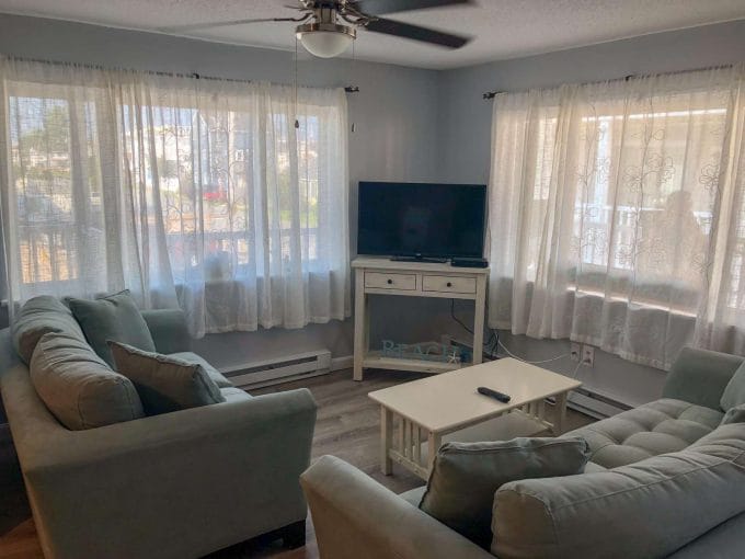 A living room with two green sofas, a white coffee table, a flat-screen TV on a console, and three large windows with sheer curtains. A ceiling fan is above.