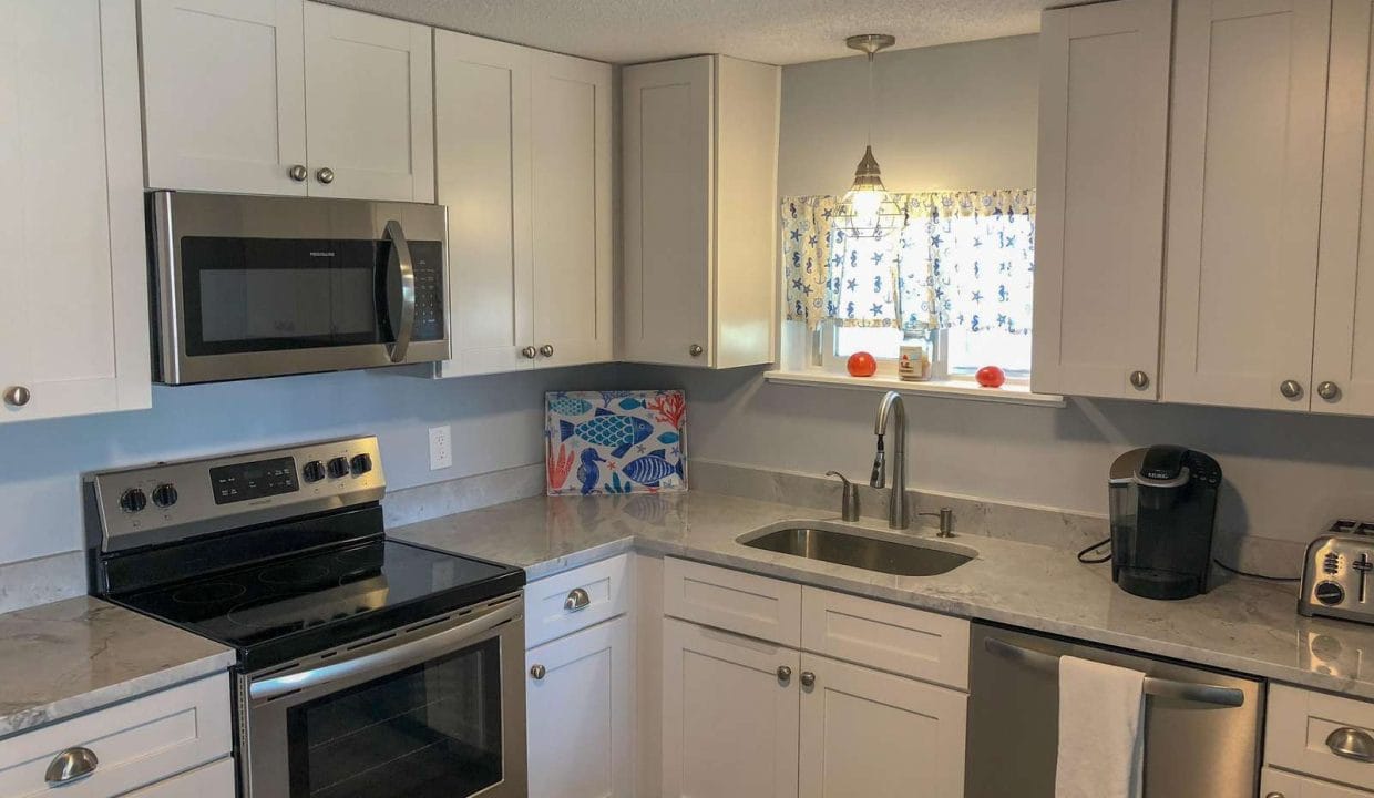 Modern kitchen with white cabinets, stainless steel appliances, a microwave, oven, dishwasher, and coffee maker. A window with floral curtains is above the sink.