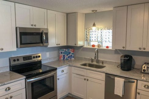 Modern kitchen with white cabinets, stainless steel appliances, a microwave, oven, dishwasher, and coffee maker. A window with floral curtains is above the sink.