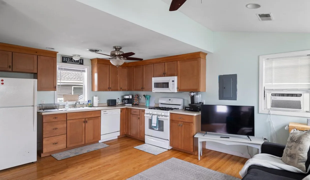 Kitchen and living area with wooden cabinets, white appliances, and a wall-mounted TV. Ceiling fans and light blue walls complete the room.