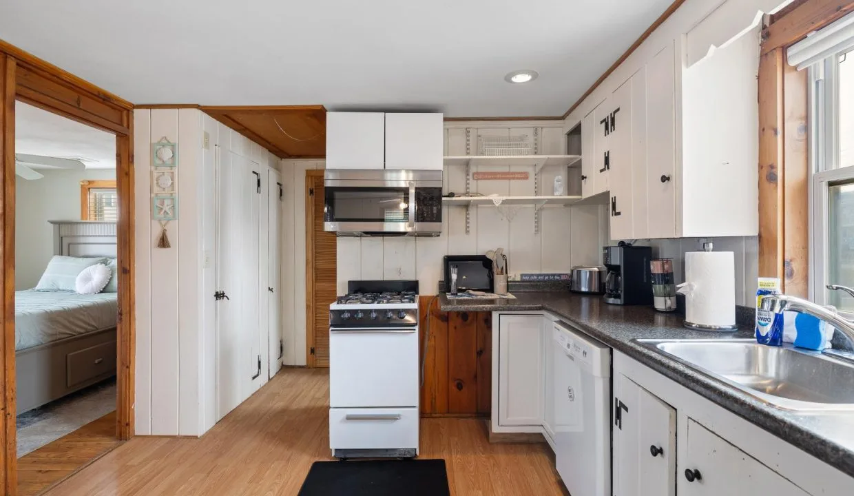 A compact kitchen with a stove, microwave, and various appliances on the counter, adjacent to a dining area. There is visible shelving with items and a doorway leading to a bedroom.