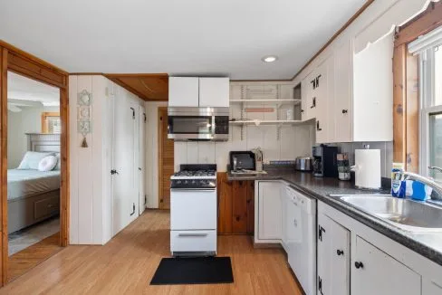 A compact kitchen with a stove, microwave, and various appliances on the counter, adjacent to a dining area. There is visible shelving with items and a doorway leading to a bedroom.