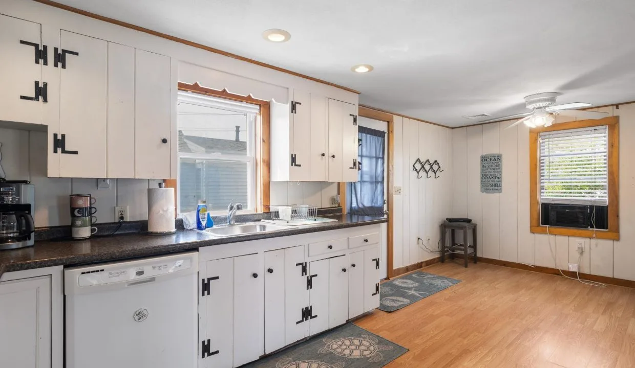 Kitchen with white cabinets, black countertops, and wooden floors. Includes a dishwasher, sink under a window, wall-mounted racks, ceiling fan, and an air conditioning unit in the window.