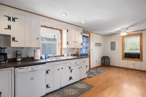 Kitchen with white cabinets, black countertops, and wooden floors. Includes a dishwasher, sink under a window, wall-mounted racks, ceiling fan, and an air conditioning unit in the window.