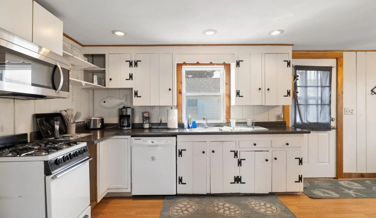 A kitchen with white cabinets, a gas stove, microwave, dishwasher, and black countertops. There is a window above the sink and a door to the right.