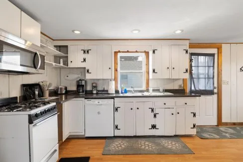 A kitchen with white cabinets, a gas stove, microwave, dishwasher, and black countertops. There is a window above the sink and a door to the right.