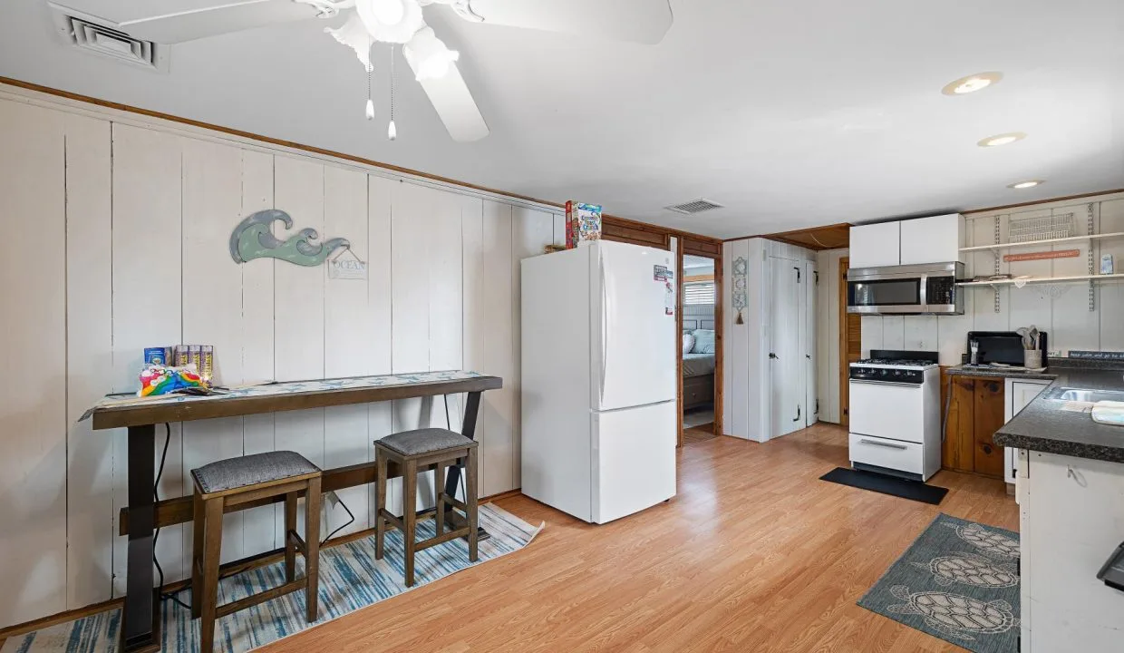 Kitchen with white walls, ceiling fan, wooden floor, table with stools, white refrigerator, and stove. Wall decor includes a wave motif. Appliances and packages are on counters and floor.