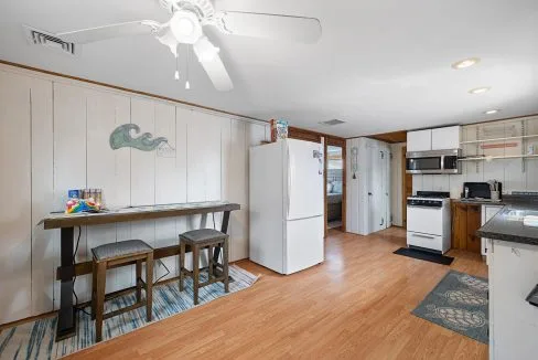 Kitchen with white walls, ceiling fan, wooden floor, table with stools, white refrigerator, and stove. Wall decor includes a wave motif. Appliances and packages are on counters and floor.