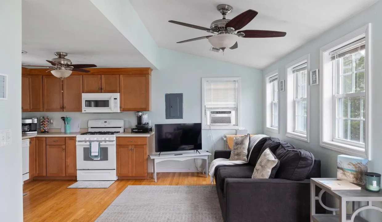 Small living area with light blue walls, wood floors, a black sofa, and a TV. Kitchen features wood cabinets, white appliances, and ceiling fans. Natural light enters through multiple windows.