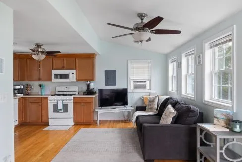 Small living area with light blue walls, wood floors, a black sofa, and a TV. Kitchen features wood cabinets, white appliances, and ceiling fans. Natural light enters through multiple windows.