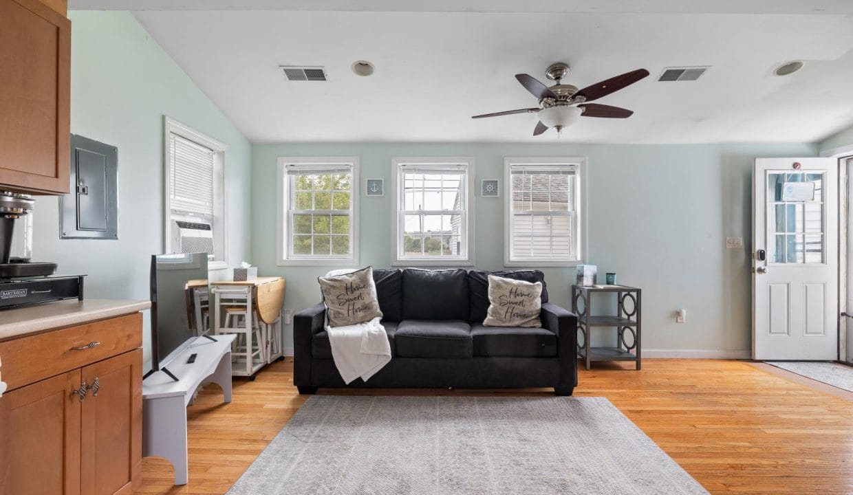 Living room with a black sofa, decorative pillows, a ceiling fan, and four windows. Light green walls and hardwood floor. A side table and a light-colored area rug are also visible.