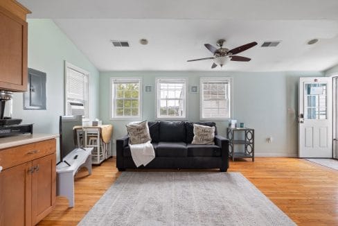 Living room with a black sofa, decorative pillows, a ceiling fan, and four windows. Light green walls and hardwood floor. A side table and a light-colored area rug are also visible.