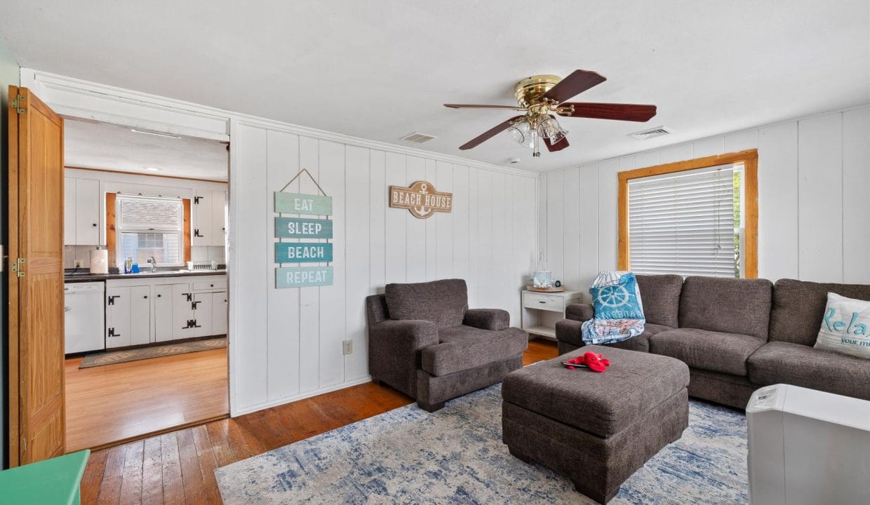 A living room with a gray sectional sofa, ottoman, and rug. Wall decor includes signs and a ceiling fan. A door opens to a kitchen with wooden cabinetry. Light fills the space from two windows.