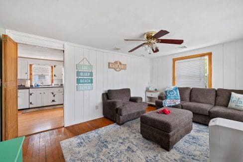 A living room with a gray sectional sofa, ottoman, and rug. Wall decor includes signs and a ceiling fan. A door opens to a kitchen with wooden cabinetry. Light fills the space from two windows.