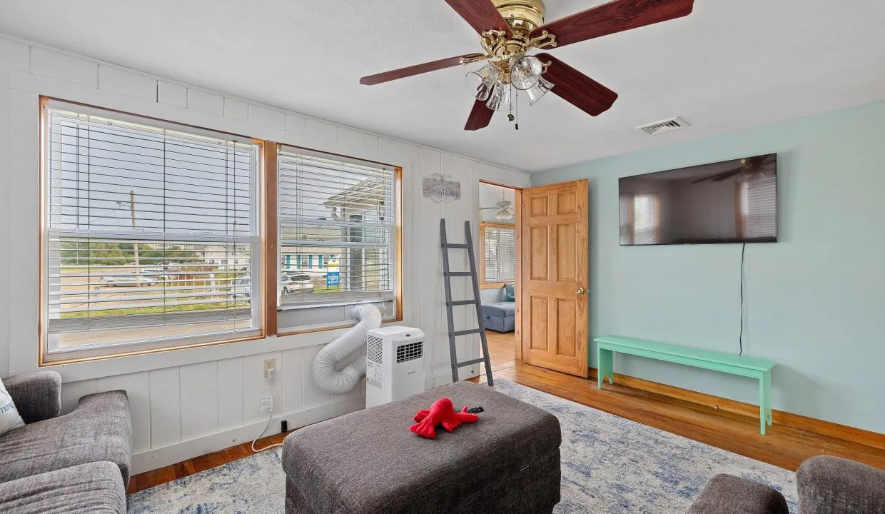 Living room with two gray sofas, a dark ottoman, wall-mounted TV, ceiling fan, light green wall, large window, and a portable air conditioner. A wooden door leads to another room.