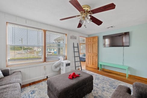 Living room with two gray sofas, a dark ottoman, wall-mounted TV, ceiling fan, light green wall, large window, and a portable air conditioner. A wooden door leads to another room.