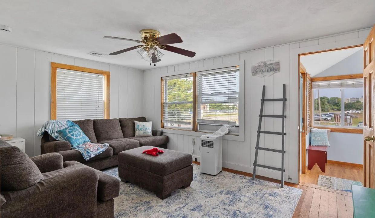 Living room with brown sectional sofa, ottoman, and patterned rug. Ceiling fan overhead, ladder shelf, and windows letting in natural light. Doorway leads to a porch area.