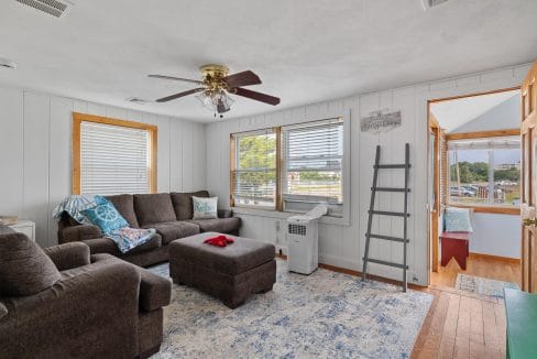 Living room with brown sectional sofa, ottoman, and patterned rug. Ceiling fan overhead, ladder shelf, and windows letting in natural light. Doorway leads to a porch area.