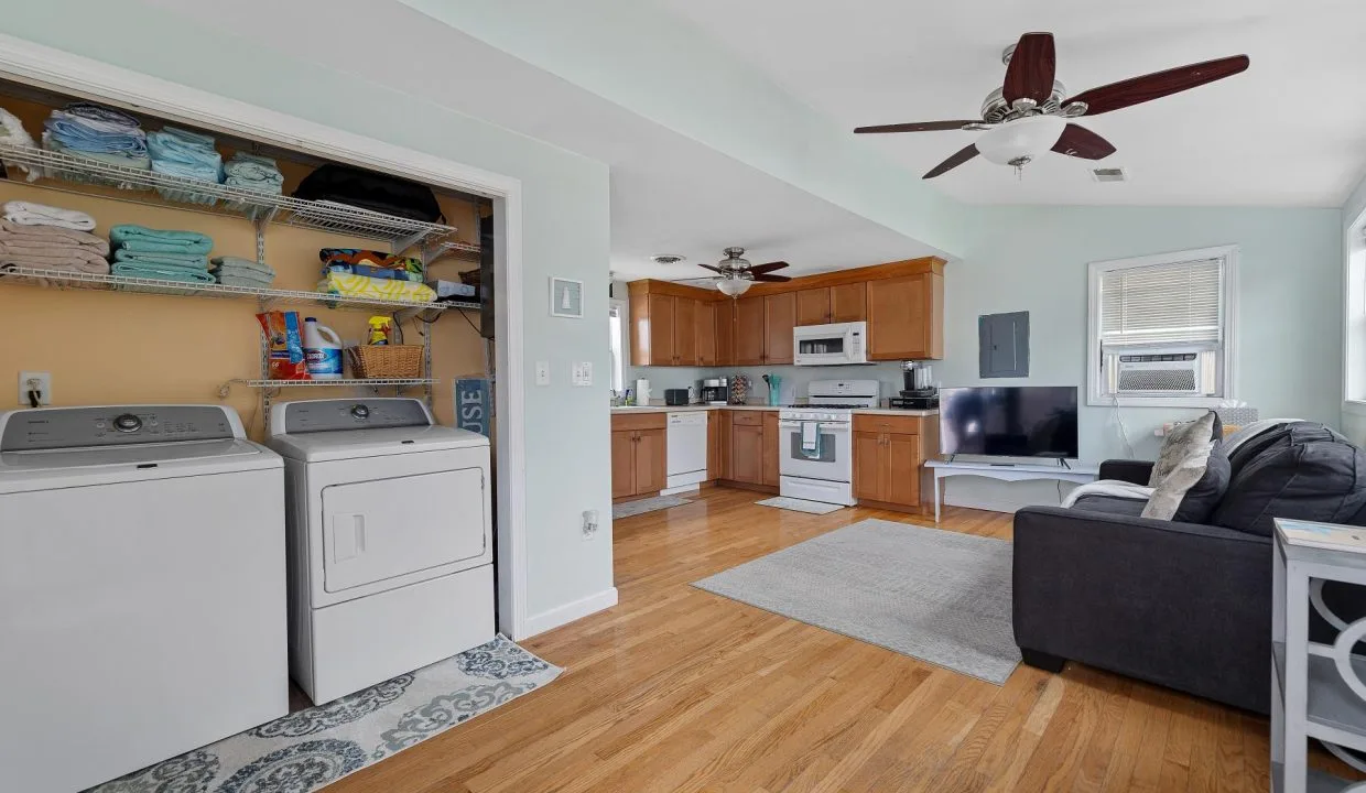 Open-plan room with a washer and dryer, shelves with towels, a kitchen area with wooden cabinets, and a living space with a couch and TV. Ceiling fans are present. Hardwood floors throughout.