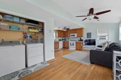 Open-plan room with a washer and dryer, shelves with towels, a kitchen area with wooden cabinets, and a living space with a couch and TV. Ceiling fans are present. Hardwood floors throughout.