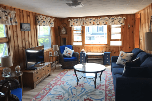 Cozy living room with wood-paneled walls, blue sofas, a round coffee table, and a TV. Nautical-themed curtains and cushions. Large windows allow natural light. Rug with floral design on the floor.