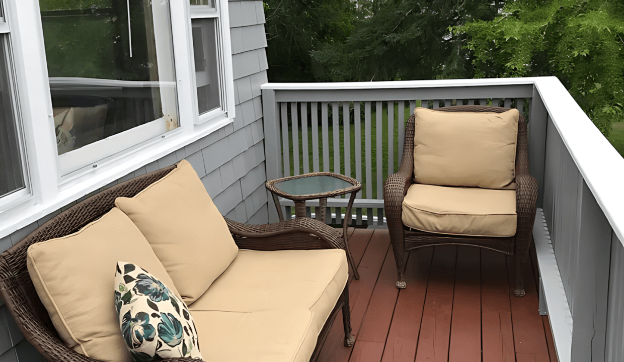 Outdoor patio with two wicker chairs and a small table on a wooden deck. A cushion with a leaf pattern rests on one chair. Gray siding and a window in the background. Trees visible beyond the railing.