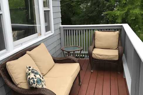 Outdoor patio with two wicker chairs and a small table on a wooden deck. A cushion with a leaf pattern rests on one chair. Gray siding and a window in the background. Trees visible beyond the railing.