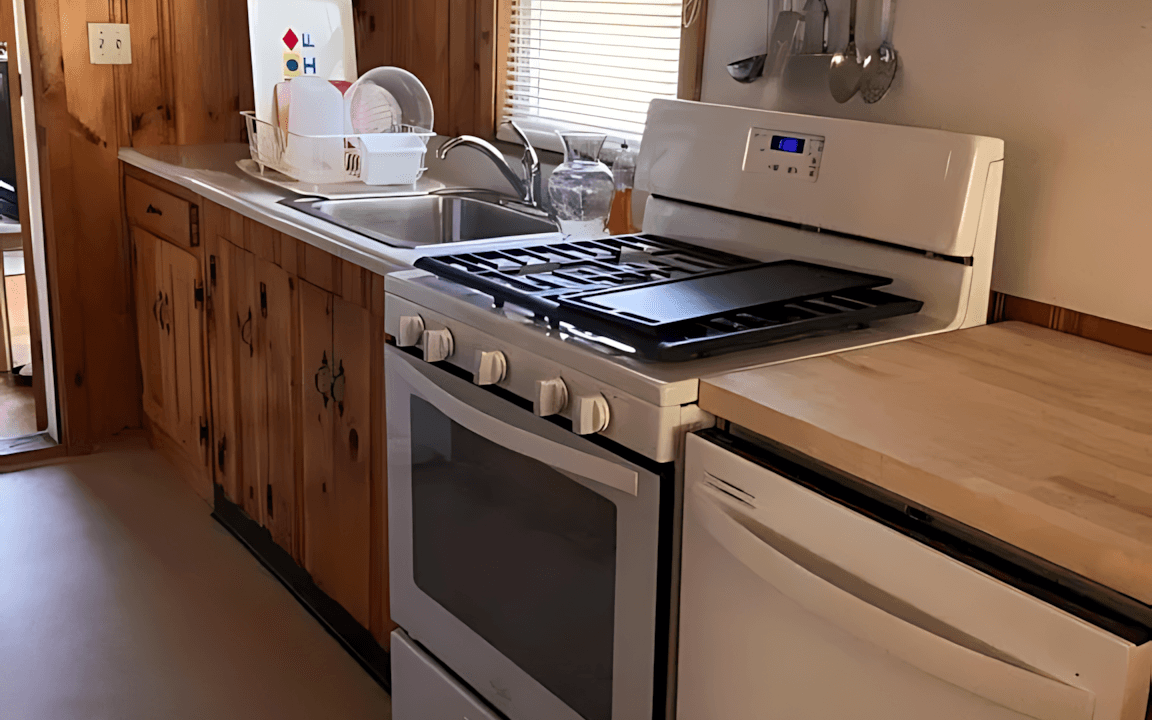 Kitchen with wooden cabinets, a stove with oven, a fridge, and utensils hanging on the wall. Dishes and a soap bottle are on the countertop near a window.