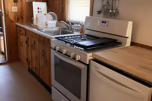 Kitchen with wooden cabinets, a stove with oven, a fridge, and utensils hanging on the wall. Dishes and a soap bottle are on the countertop near a window.