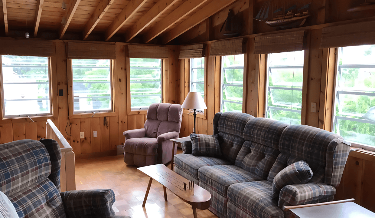 A cozy sunroom with wooden walls, featuring plaid sofas, a recliner, a ceiling fan, and a small wooden table, with large windows offering a view of greenery outside.