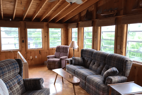 A cozy sunroom with wooden walls, featuring plaid sofas, a recliner, a ceiling fan, and a small wooden table, with large windows offering a view of greenery outside.