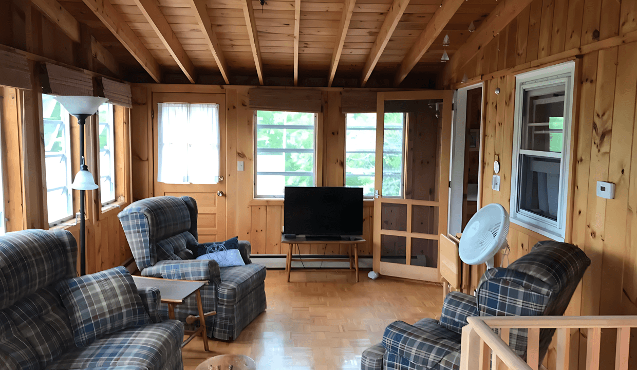 A cozy wooden living room features plaid armchairs, a coffee table, and a flat-screen TV. The room has large windows, a ceiling fan, and parquet flooring, offering a rustic and comfortable ambiance.