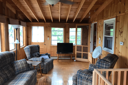 A cozy wooden living room features plaid armchairs, a coffee table, and a flat-screen TV. The room has large windows, a ceiling fan, and parquet flooring, offering a rustic and comfortable ambiance.