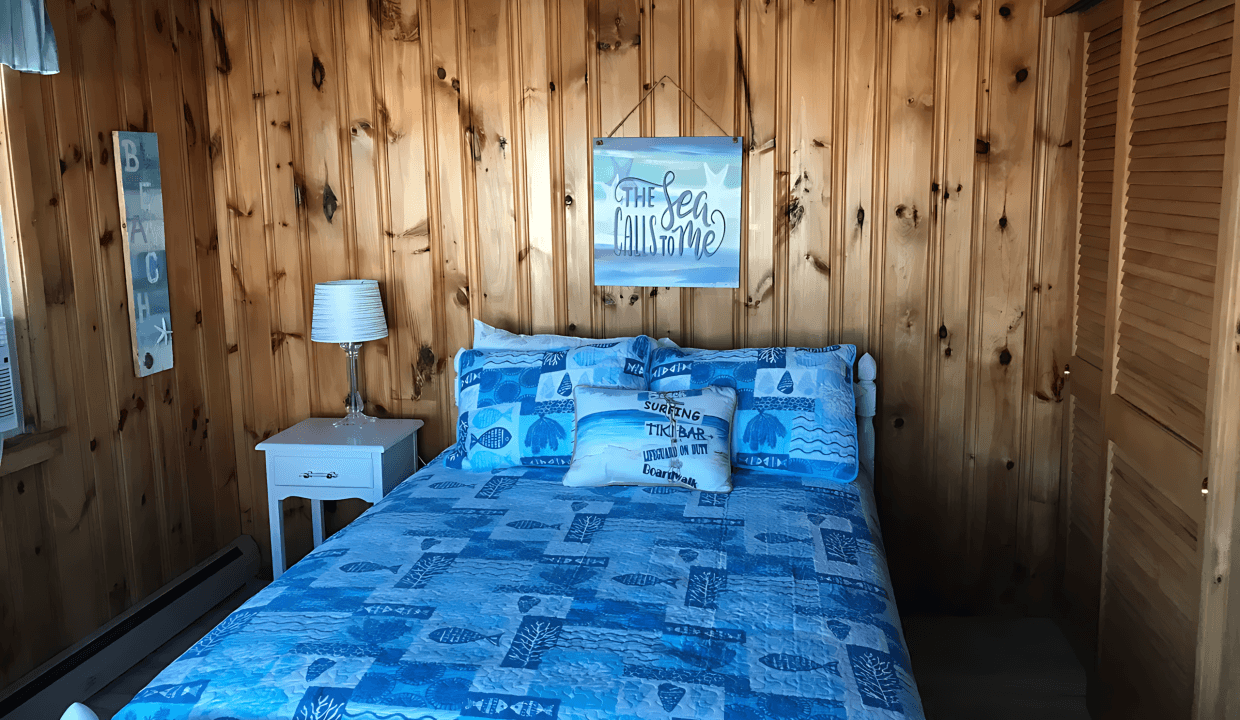 Bedroom with wood-paneled walls featuring a bed with blue patterned bedding, a side table with a lamp, and a wall art piece above the bed.